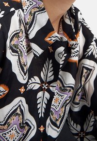 Camisa de tela negra con un audaz patrón geométrico colorido, con acentos en blanco, naranja y púrpura en el cuello y las mangas.