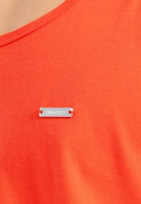 Pomarańczowa koszulka z materiału o gładkiej fakturze, z metaliczną srebrną metką z wygrawerowanym napisem "SMILODOX" przymocowaną na klatce piersiowej.