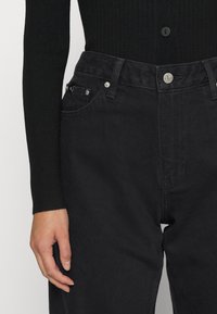 Torso inferior vestido con pantalones vaqueros negros de cintura alta y una blusa de manga larga de canalé negra, con la mano izquierda relajada al lado.