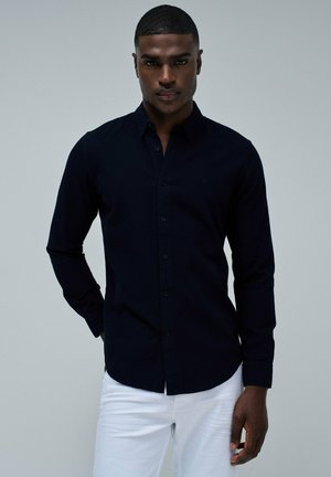 Navy blue button-up shirt with long sleeves, textured fabric, and a classic collar, paired with white pants. No visible logos or patterns.