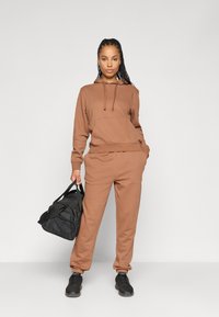 Brun hettegenser og sweatpants-sett laget av mykt materiale, med en frontlomme og elastiske mansjetter. Modellen holder en svart treningsbag.