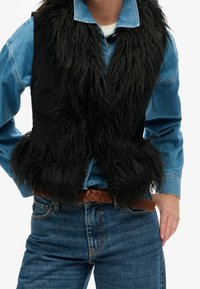 Svart faux fur-väst med en lurvig textur, lagd över en blå denimskjorta. Har en figurnära passform och ett brunt flätat bälte i midjan.