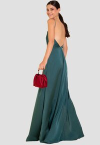 Abito lungo in satin verde acqua con profondo scollo sulla schiena, spalline sottili e silhouette fluida; abbinato a una piccola borsa rossa arricciata.