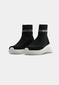 Sapatilhas slip-on de malha preta com uma superfície texturizada, apresentando uma sola de borracha branca e uma faixa de logotipo cinza no tornozelo.