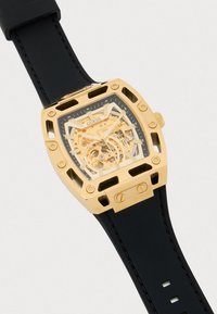 Relógio esqueleto dourado com bracelete de borracha preta. Caixa quadrada com recortes, exibindo engrenagens intrincadas sob um vidro. Logotipo exibido.