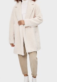 Světlý béžový, měkký, oversized kabát s jemným texturovaným povrchem, širokými klopami a otevřeným předním dílem. Nesený přes přiléhavý rolák a béžové kalhoty.
