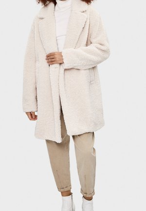 Manteau surdimensionné en beige clair, en peluche, avec une surface douce et texturée, des revers larges et une ouverture frontale. Porté sur un col roulé ajusté et un pantalon beige.