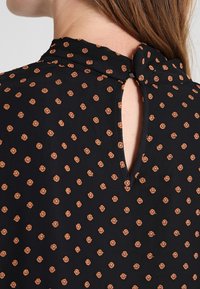 Close-up of a woman’s neck and upper back wearing a black top with small orange floral dots and a keyhole button closure.