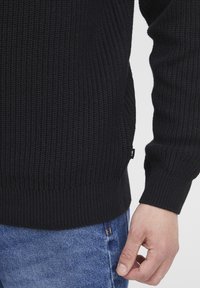 Black knitted sweater with ribbed cuffs and hem, featuring a side slit and a small brand tag. Worn over blue denim jeans.