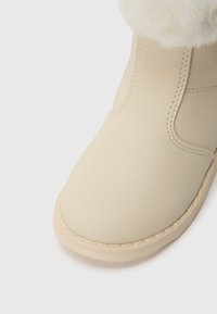 Botte d'hiver beige avec une tige en cuir lisse, des coutures visibles, un bout arrondi et une doublure en fourrure blanche et duveteuse visible en haut.
