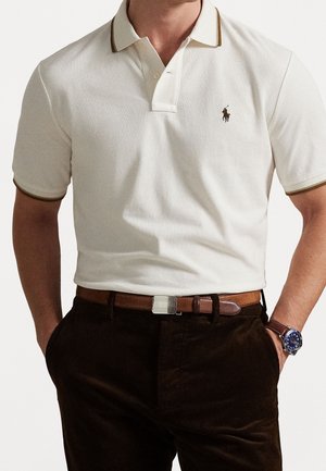 Polo shirt blanc avec accent marron sur le col, arborant un petit logo. Associé à un pantalon en velours côtelé marron et une ceinture marron. Montre au poignet.