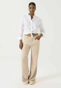 Camisa blanca de botones atada en la cintura, combinada con pantalones de pierna ancha beige. El modelo lleva zapatos claros; el fondo es blanco liso.