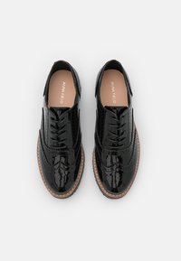 Scarpe stringate in pelle verniciata nera con design brogue, caratterizzate da una texture liscia e una suola color marrone chiaro. Branding minimo sulla soletta.