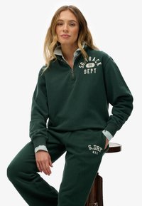 Felpa verde scuro con collo a zip e scritta bianca. Abbinata a pantaloni della tuta coordinati con polsino a righe e dettaglio logo. Materiale morbido.