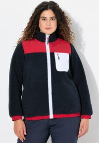 Veste en polaire bleu marine avec une partie supérieure rouge, fermeture éclair blanche à l'avant et poche. Matériau texturé avec des poignets et un ourlet élastiques.