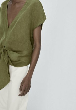 Personne portant un chemisier court olive noué et un pantalon blanc sur un fond uni gris clair.