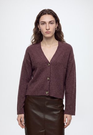 Femme aux cheveux bruns ondulés portant des boucles d'oreilles créoles en or, un cardigan bordeaux boutonné et une jupe en cuir marron foncé sur un fond uni.