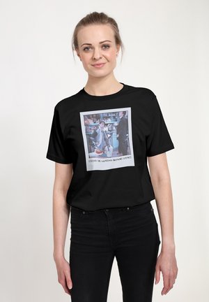 Kvinde med lyst hår sat op iført en sort skjorte med et farverigt trykt foto af to personer i et køkkenmiljø.