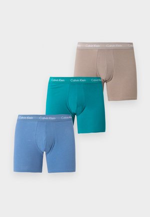 Trois paires de boxers pour hommes Calvin Klein en beige, sarcelle et bleu, disposées en quinconce sur un fond blanc.