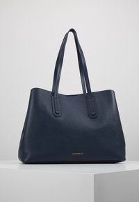 Granatowa skórzana torba na ramię o teksturowanej powierzchni, z dwoma długimi uchwytami i złotym wytłoczonym logo z przodu. Minimalistyczny design i otwarty górny zamek.