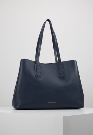 Navy-nahka-tote laukku, jossa on teksturoitu pinta, kaksi pitkää kahvaa ja kultaa kohokuvioitu logo edessä. Minimalistinen muotoilu ja avoin yläosa.