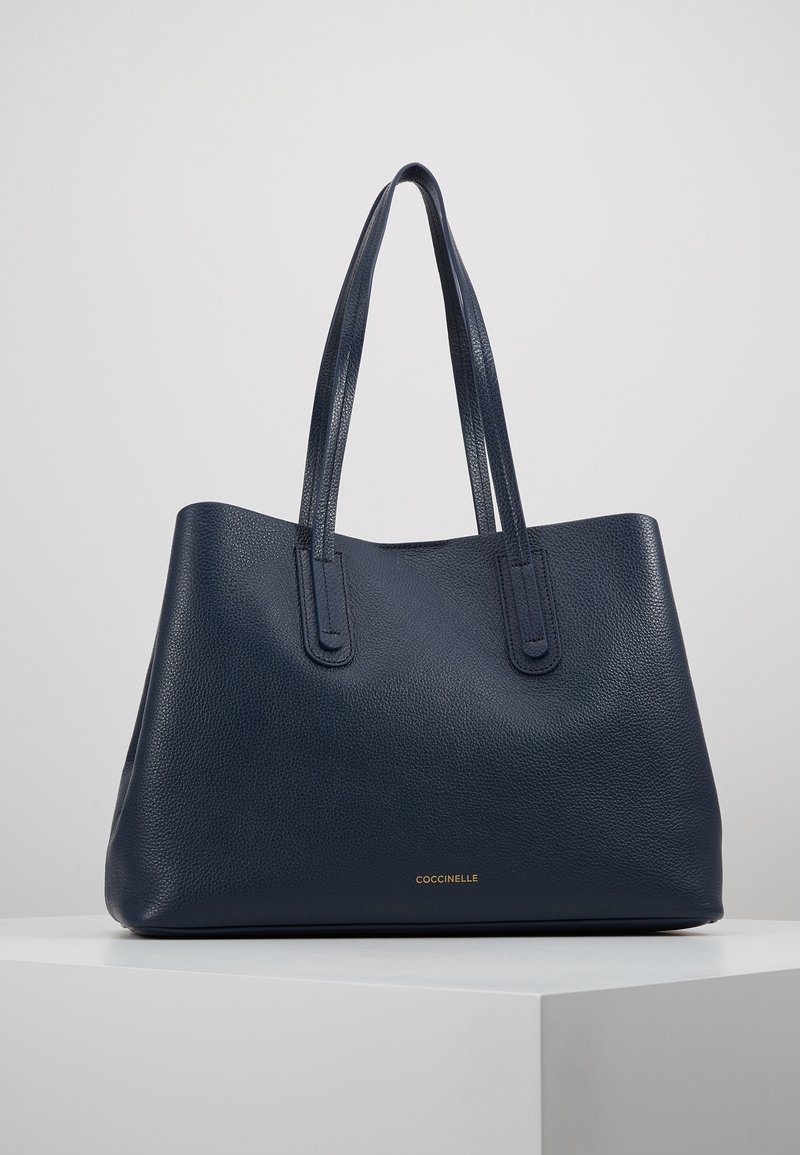 Granatowa skórzana torba na ramię o teksturowanej powierzchni, z dwoma długimi uchwytami i złotym wytłoczonym logo z przodu. Minimalistyczny design i otwarty górny zamek.