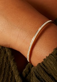 Gouden armband met een slank ontwerp, voorzien van ingelegde heldere kristallen langs één rand, tegen een getextureerd huidoppervlak.