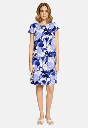 Vrouw staand met een blauw-wit bloemen kort mouw top en bijpassende knielange rok met witte sandalen op een neutrale achtergrond.