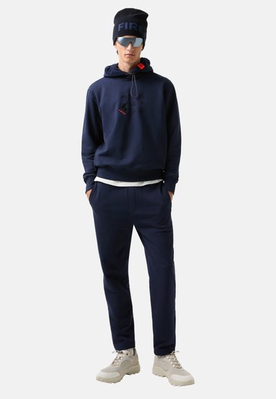 Muž v námořnickém hoodie s grafickými detaily, odpovídajících námořnických kalhotách, béžových teniskách, tmavé čepici s nápisem a reflexních slunečních brýlích, stojící s rukama v kapsách.