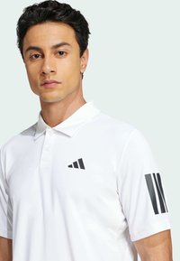 Hvit polo-skjorte laget av pustende stoff med svart logo på brystet og stripete detaljer på ermene. Glatt tekstur, normal passform.