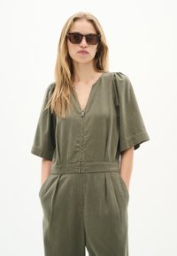 Salopetă verde măsliniu cu mâneci scurte și voluminoase, fermoar frontal și buzunare laterale. Material neted, ușor, cu o croială lejeră.