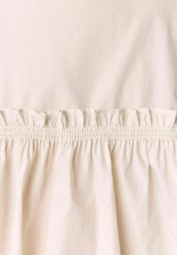 Blouse beige clair avec une texture froncée et un ourlet volanté. Tissu lisse avec un subtil éclat, mettant en valeur son design aérien.