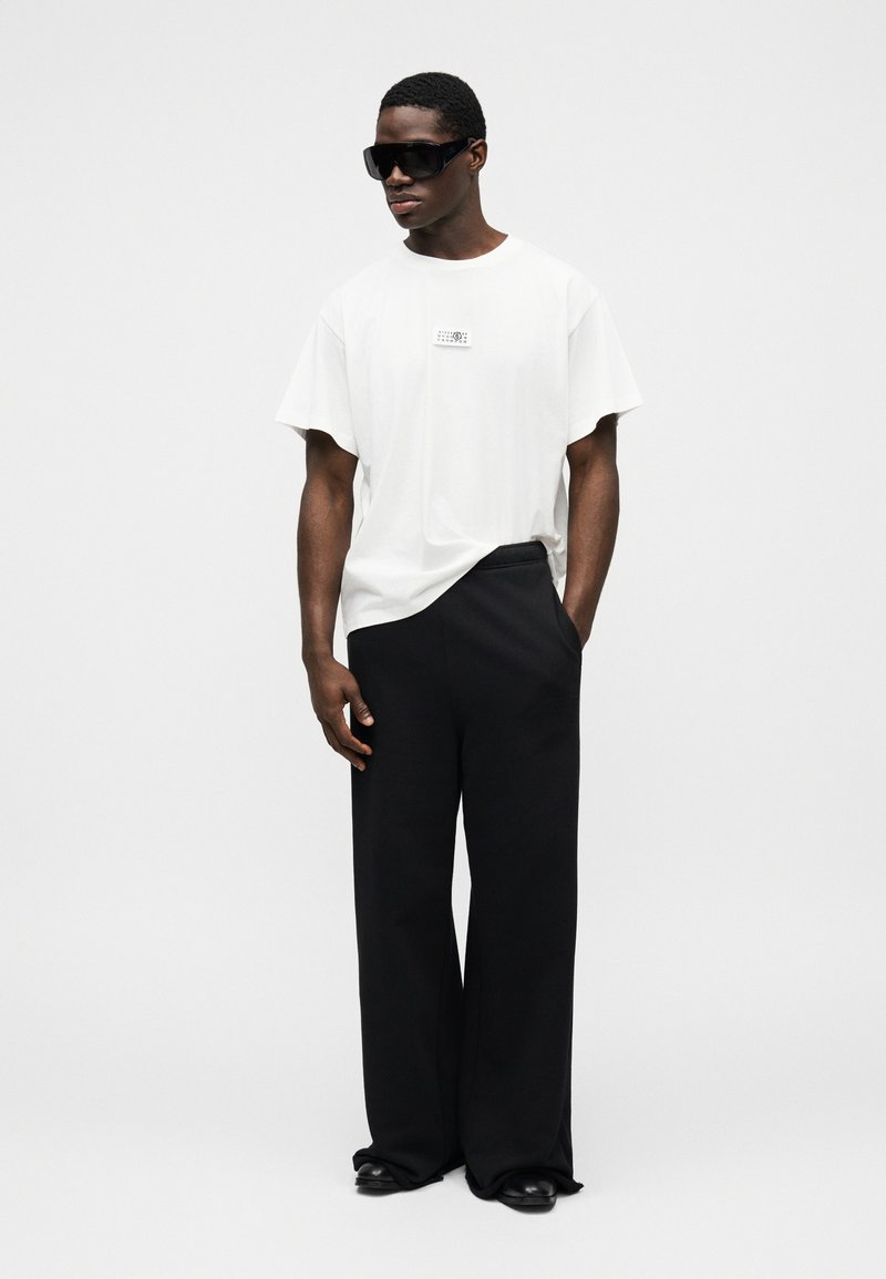 Hvid oversized t-shirt med grafik på brystet, kombineret med løse sorte bukser. Tilbehør inkluderer mørke solbriller og sorte sko.