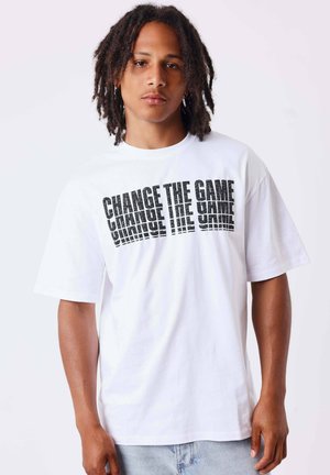 Giovane uomo con dreadlock di media lunghezza che indossa una t-shirt bianca con "CHANGE THE GAME" stampato in lettere nere grassetto e distorte.