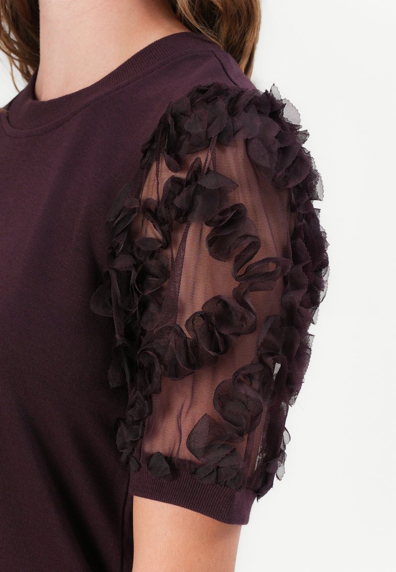 T-shirt viola con scollatura a girocollo testurizzata e maniche trasparenti in rete nera a volant, caratterizzata da motivi floreali e una silhouette aderente.