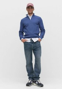 Blå stickad tröja med dragkedja fram, bärs över en vit skjorta. Tillsammans med ljusa jeans och svarta sneakers med vita detaljer.