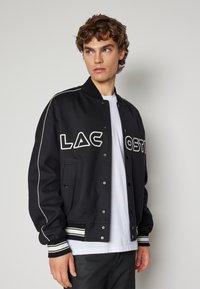 Svart varsityjacka med vita ackcenter, ribbad mudd på ärmarna och knappfront. Har stor "LACOSTE"-logotyp på bröstet. Vadderad textur.