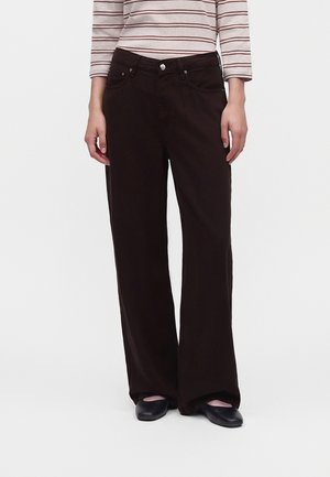 Modelo con pantalones marrón oscuro de pernera ancha, una camiseta de manga larga a rayas y zapatos planos negros, sobre un fondo blanco liso.