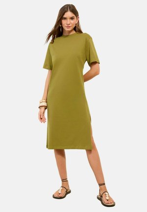 Žena nosí oličkovo zelené tričkové šaty po kolená s krátkymi rukávmi, hnedé sandále, zlaté kruhové náušnice a masívne náramky.