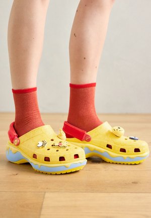 Persoon draagt gele kindersabots met bijen- en honingraatdecoraties, rode bandjes en rode sokken, staand op een houten vloer.