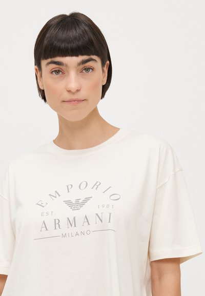 Bež majica od pamuka s kratkim rukavima, sa središnjim sivim ispisanim logotipom koji uključuje "EMPORIO ARMANI" i "MILANO", te godinu "1981."