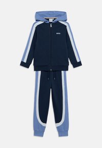 Σκούρο μπλε ναυτικό και ανοιχτό μπλε φόρμα με ζακέτα με κουκούλα και φερμουάρ, ρίγες στους ώμους και αντίστοιχο παντελόνι με στενά πόδια και ελαστική μέση.