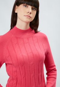 Gestrickter Pullover in leuchtendem Pink. Verfügt über einen hohen Rippenkragen und ein strukturiertes Zopfmuster auf der Vorderseite mit langen Ärmeln.