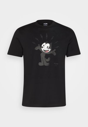 Uomo con maglietta nera con grafica di Felix the Cat e pantaloni di pelle blu, in piedi davanti a uno sfondo chiaro e semplice.