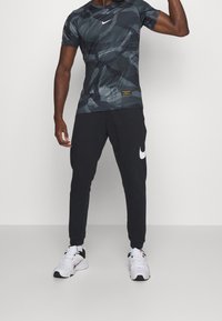 Conjunto deportivo de Nike que incluye una camiseta de manga corta con un patrón de camuflaje oscuro y pantalones de chándal negros ajustados con un logo blanco. Zapatillas blancas.