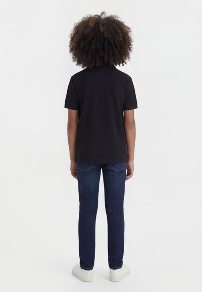 Enfant aux cheveux bouclés portant un t-shirt noir à manches courtes, un jean bleu et des baskets blanches, debout de dos sur un fond uni.