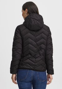 Črna puhovka z vzorcem šivov v obliki črke V, priložena kapuca in elastični manšeti, kombinirana s temnimi modrimi kavbojkami.