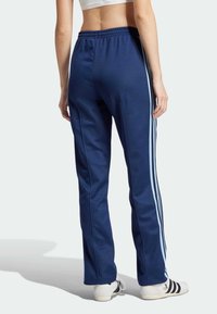 Osoba oblečená v tmavomodrých teplákoch so svetlomodrými pruhmi na bokoch a bielymi teniskami, zobrazená zozadu na jednoduchom pozadí.