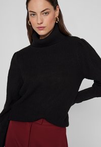 Melnas turtleneck džemperis ar ribotu tekstūru, ar garām piedurknēm un brīvu piegriezumu, apvienots ar dziļi sarkanu apģērbu apakšā.