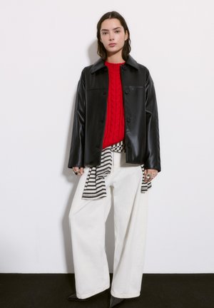 Zwarte leren jas met knopen aan de voorkant, rode kabelgebreide trui, wijde witte broek met een gestreepte tailleband en puntige zwarte schoenen.